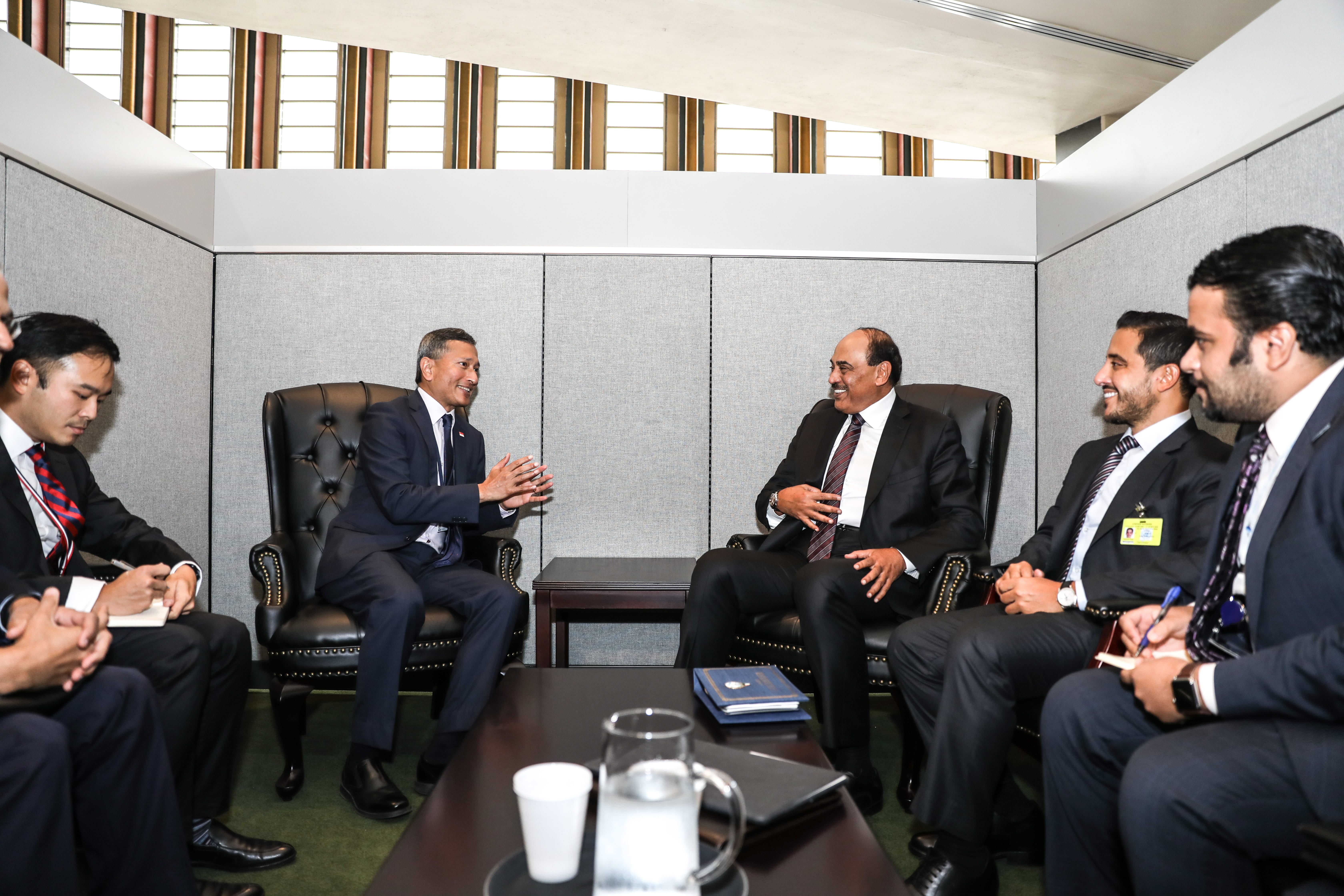 Group of men in suits seated in a meeting room with patterned walls and unique window architecture.